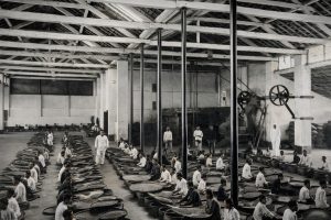 Foto hitam-putih hasil restorasi yang menampilkan suasana pabrik pengolahan kopi pada awal abad ke-20. Puluhan pekerja duduk berbaris rapi di dalam bangunan besar beratap kayu, memilah kopi menggunakan tampah besar.