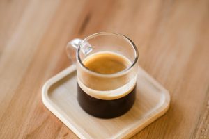 Close-up kucuran espresso base kental berwarna cokelat gelap dengan lapisan crema keemasan yang tebal, mengalir lembut ke dalam cangkir kecil di atas meja kayu.